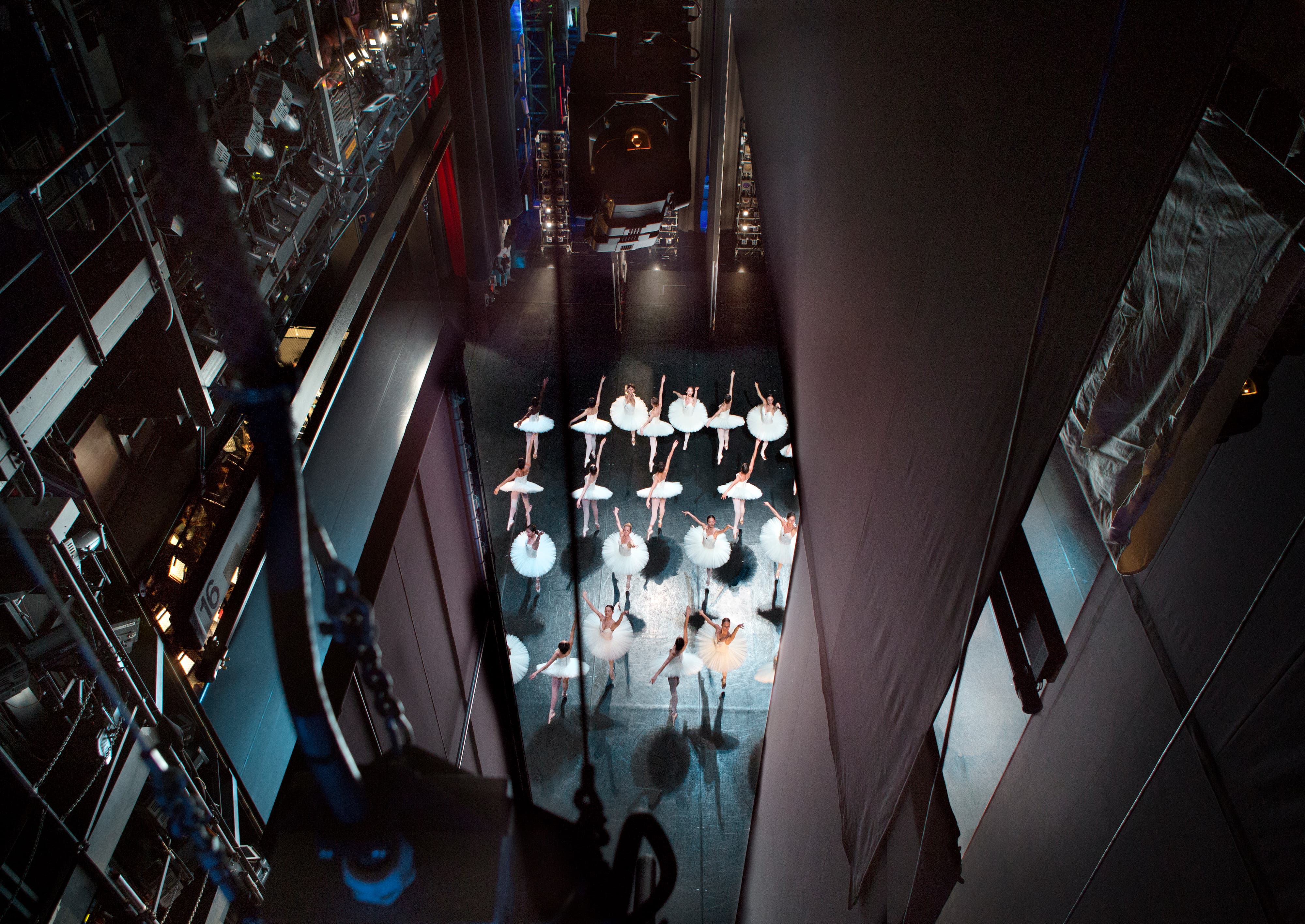 Bovenzicht van een groep ballerina’s in witte tutu’s op het toneel, gefotografeerd van hoog in de toneeltoren tussen lichtbruggen en technische installaties.